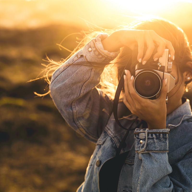 Femme en veste jean photographiant avec un appareil photo vintage au coucher de soleil, lumière dorée et contre-jour.