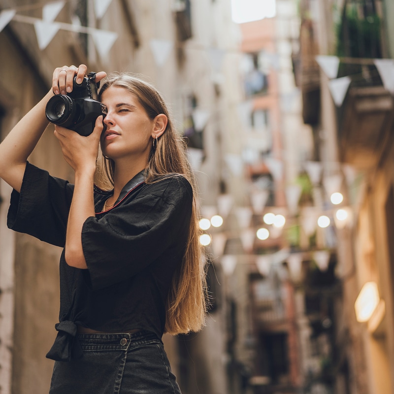 Jeune femme blonde prenant une photo avec un appareil noir dans une ruelle urbaine. Des drapeaux triangulaires décorent la rue.