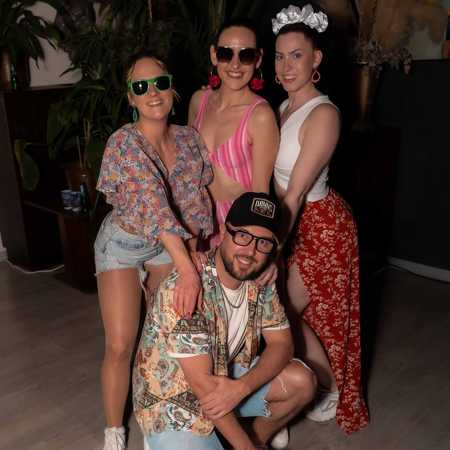 Quatre amis souriants, en tenues estivales colorées. Un homme accroupi, casquette DJINNS, et trois femmes en robes, shorts et maillot de bain. Ambiance joyeuse.
