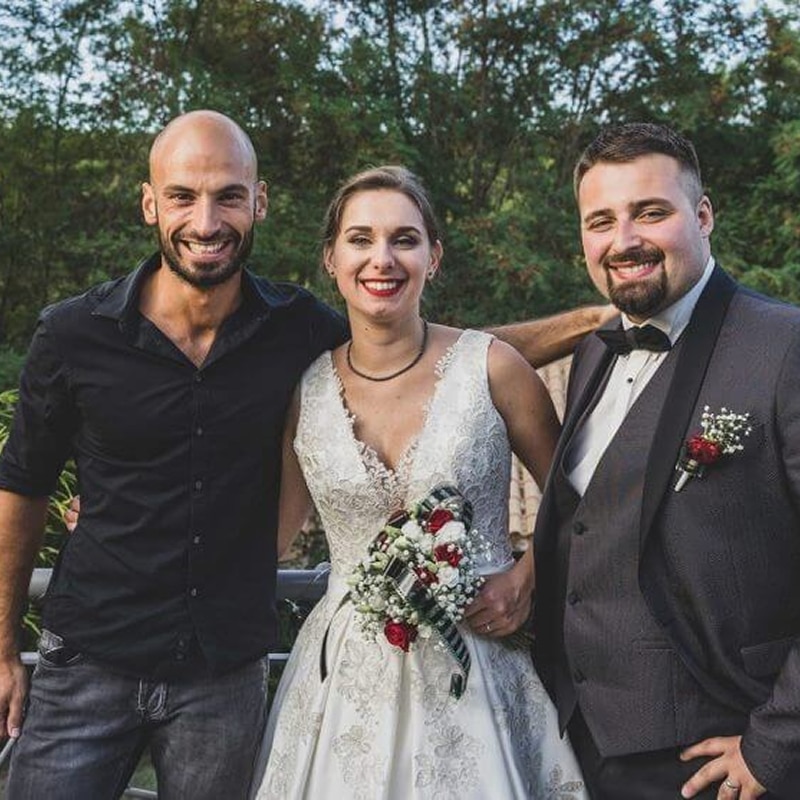 Trois personnes souriantes: une mariée avec bouquet, un marié en costume et un ami. Scène joyeuse en extérieur.