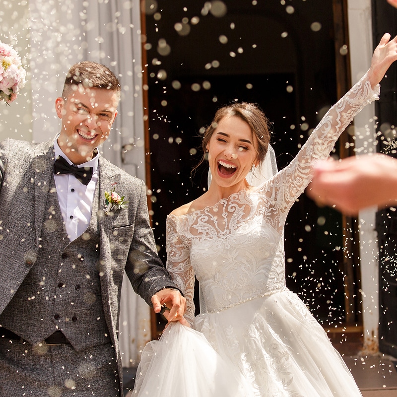Mariés souriants et joyeux sortant d'une église sous une pluie de confettis. La mariée en robe blanche lève le bras, le marié en costume gris la tient.