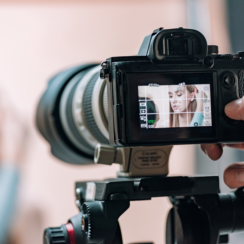 Appareil photo professionnel sur trépied, écran affichant une femme en gros plan et réglages (MF, STBY, 0:00). Tournage vidéo.