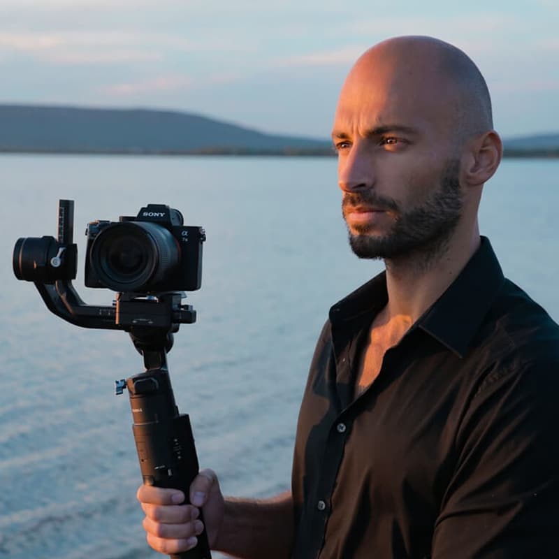Photographe ou vidéaste concentré tenant une caméra Sony α7c sur gimbal, avec un lac et montagnes au coucher du soleil.